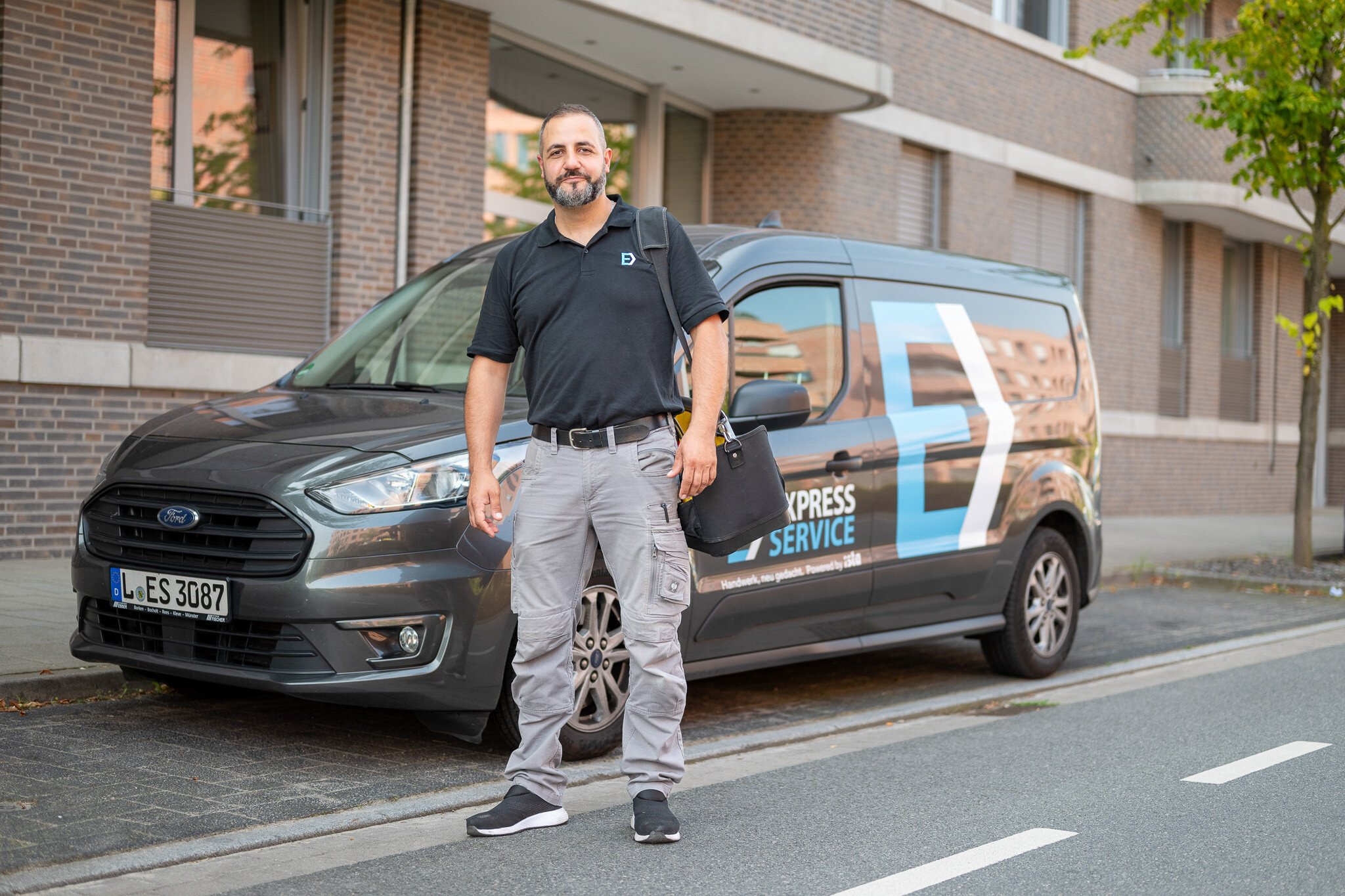 Servicetechniker mit Werkzeugtasche steht einsatzbereit vor seinem ista Express Service Servicefahrzeug vor Wohngebäuden